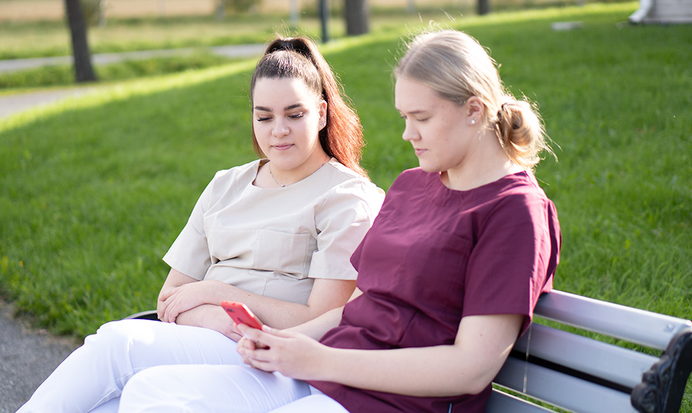 Kaksi nuorta hoitajien vaatteissa istuu ulkona penkillä ja selaa puhelinta.